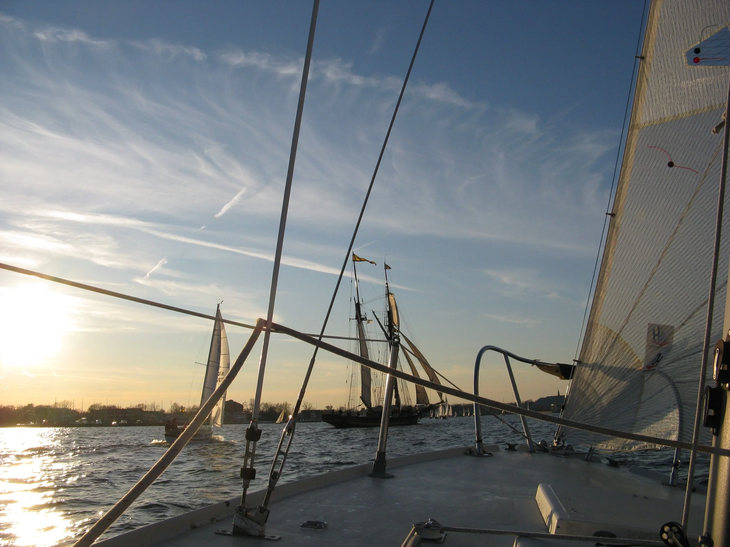 View from a sailboat during sunset, showing other sailboats and a distant shoreline with houses under a partly cloudy sky.