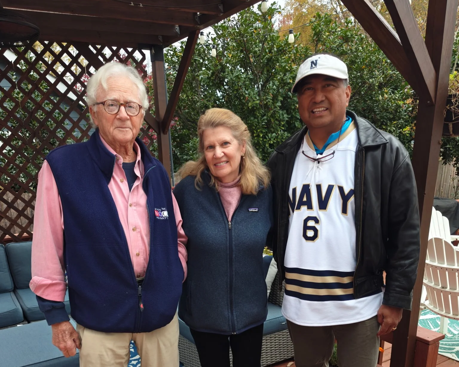 Three people standing outdoors in a backyard, posing for the camera. An elderly man on the left, a middle-aged woman in the center, and a middle-aged man on the right wearing a navy jersey and a cap.
