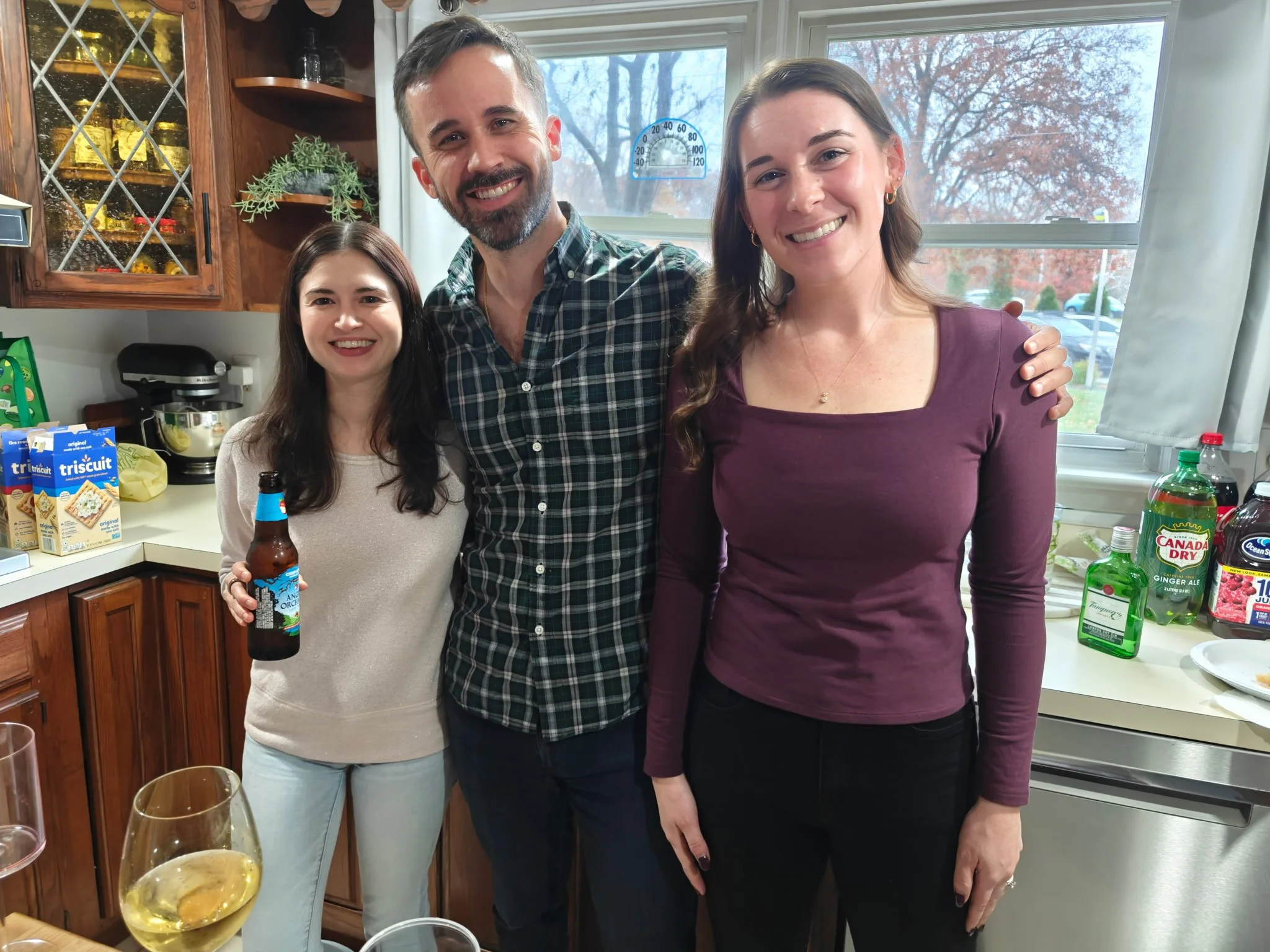Three people standing in a kitchen, smiling at the camera. One woman is holding a bottle of beer. The kitchen has a window in the background, with various drinks and kitchen items visible.