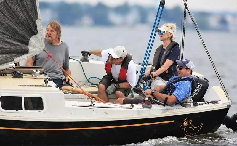 Four people on a sailboat with a black and white hull, sailing on a lake or river with trees and water in the background.