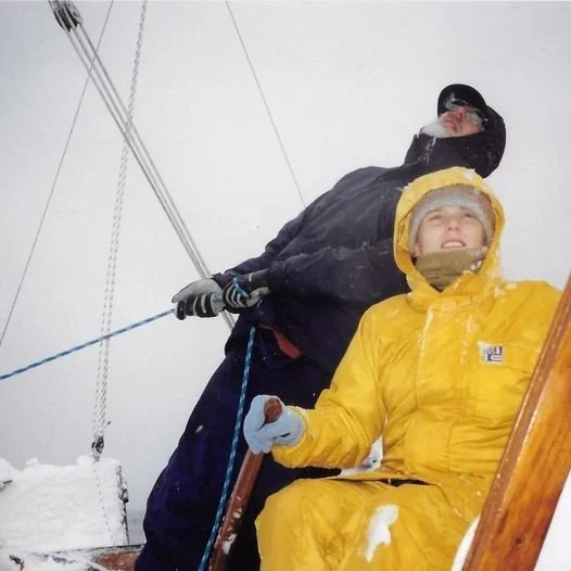 Two people in winter gear, one in a yellow jacket and the other in a black jacket, on a snowy mountain with ski equipment, under a cloudy sky.