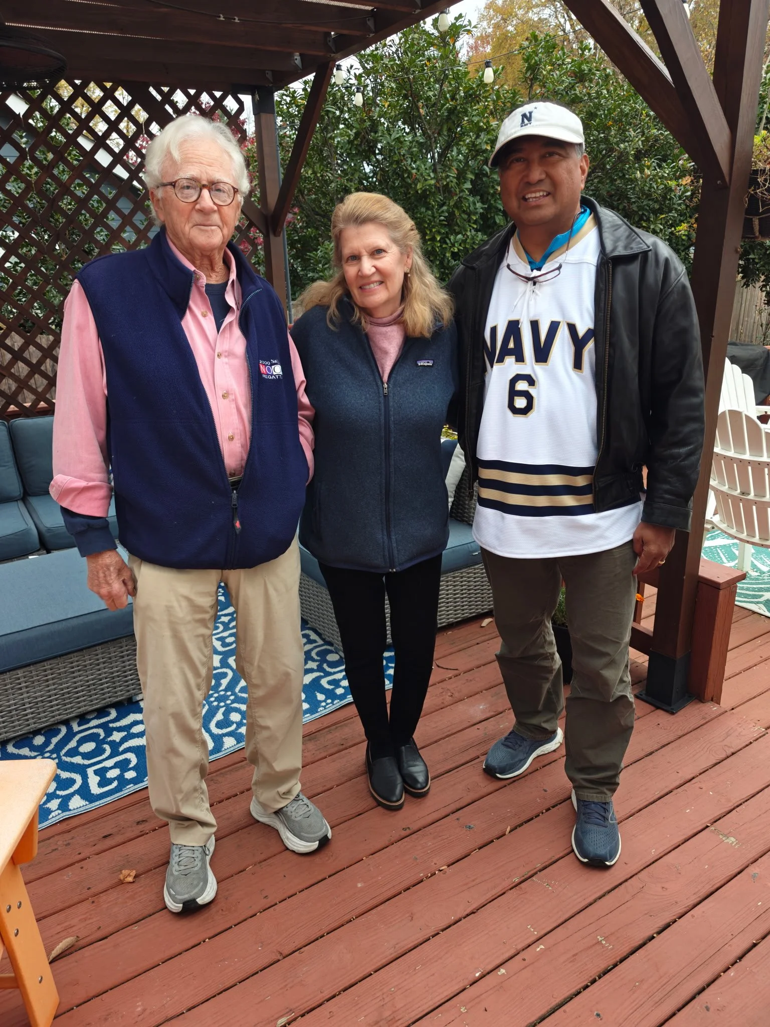 Three people standing on a wooden deck outdoors, smiling at the camera. They are dressed casually, with a man wearing a navy jersey, a woman in a blue vest, and another man in a pink shirt and navy vest.