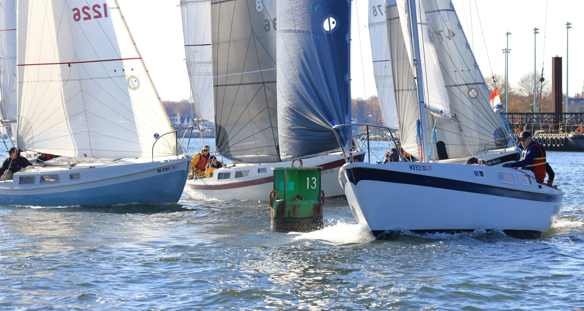Sailboats racing in a regatta with a green marker buoy numbered 13 in the water, and several crew members actively managing their sails.