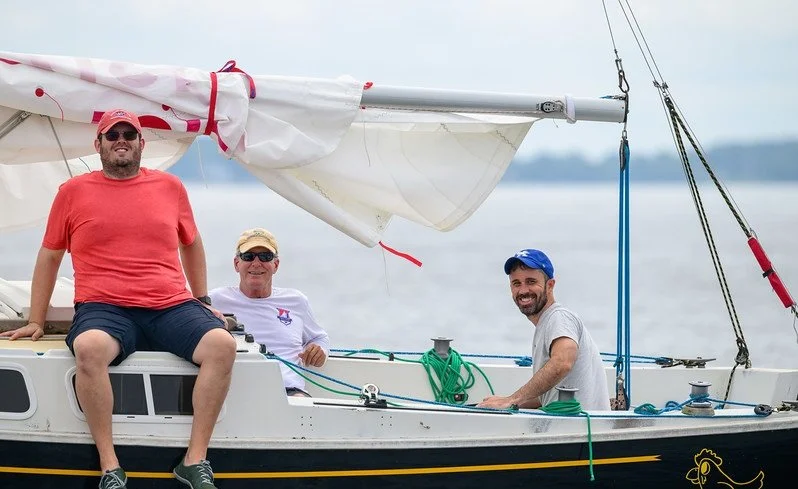 Three men on a sailboat smiling, with water and a cloudy sky in the background.