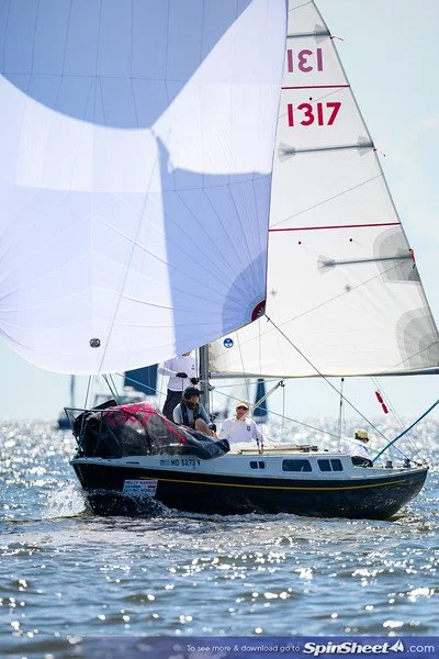 A sailboat with large sails sailing on water with sunlight reflecting off the surface, and two people on board.