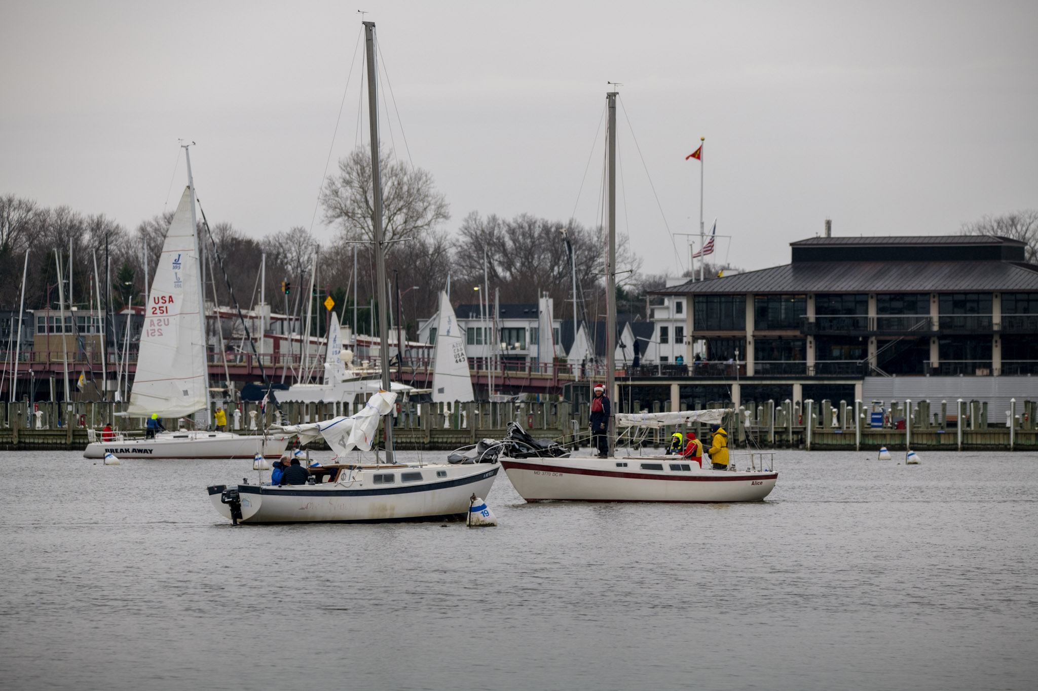 Sailboats on a calm body of water with people on board, a pier with buildings and more sailboats in the background, and an overcast sky.