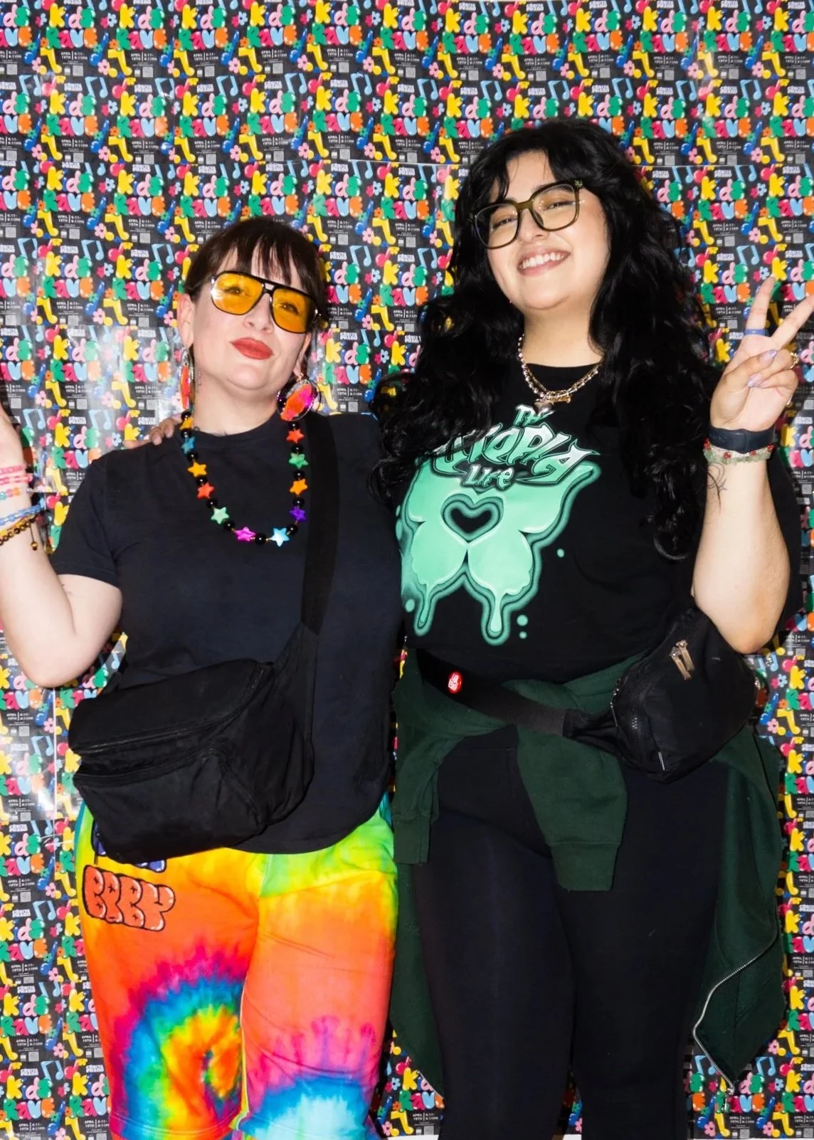 Two women smiling and posing in front of a colorful, patterned background. They are wearing vibrant, casual clothing with accessories.