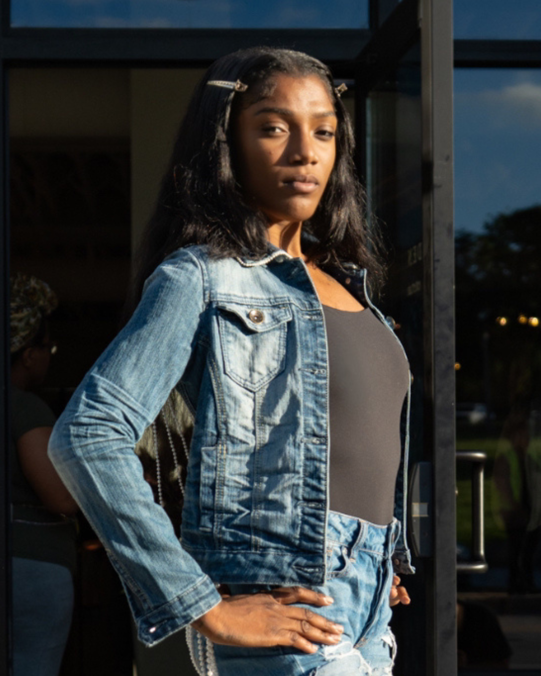 Young woman in a denim jacket and black top standing outside at dusk.