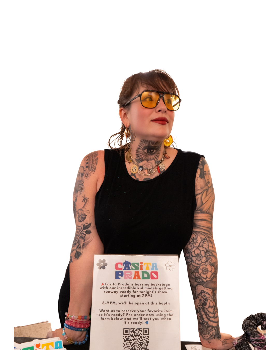 Tattooed woman with blonde hair, wearing yellow glasses and gold jewelry, standing behind a booth with a sign, at night. She has tattoos on her arms and neck, and is wearing a black sleeveless top.
