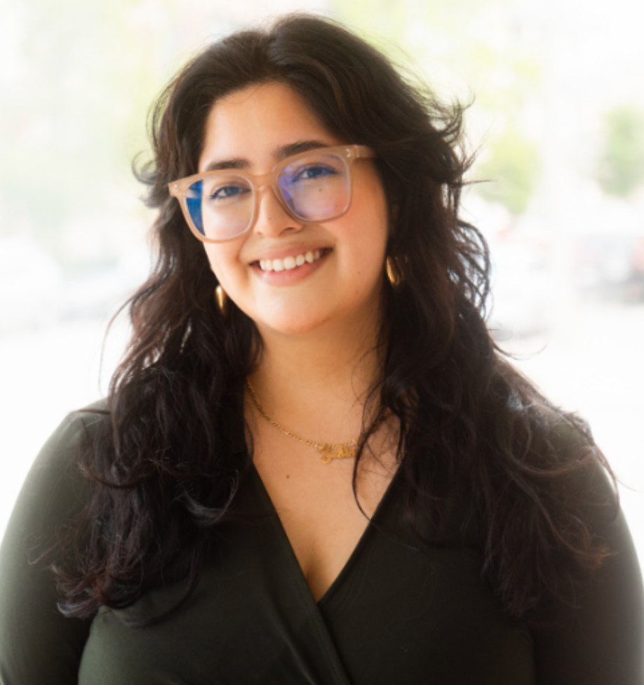 A woman with curly dark hair, wearing glasses, gold jewelry, a black top, smiling in front of a bright window.