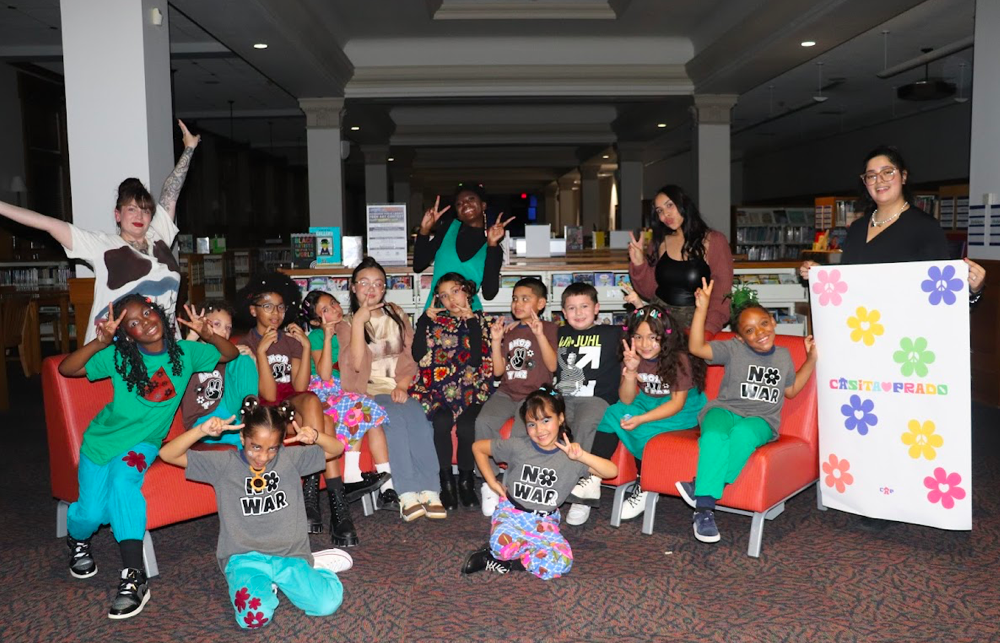 Group of children and adults posing for a photo in a library. The children are sitting and standing, making peace signs. One adult is holding a large colorful poster with flowers and the words 'Castillo Prado.' The background shows bookshelves and a quiet library setting.