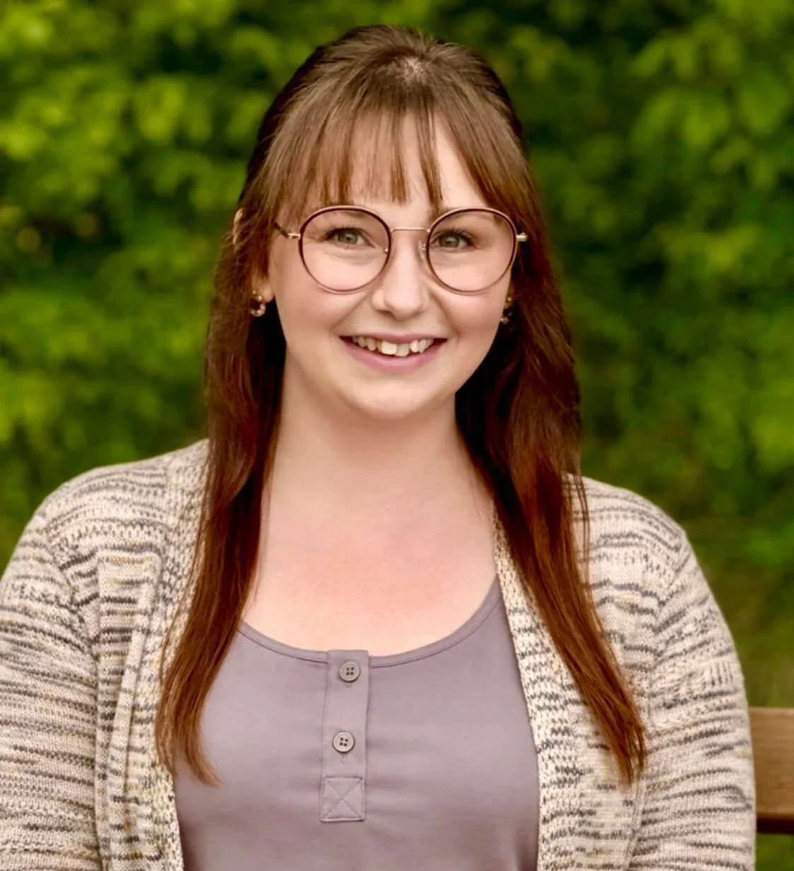 A smiling woman with red hair, glasses, and earrings, wearing a beige patterned cardigan and a gray top, standing outdoors with green foliage in the background.