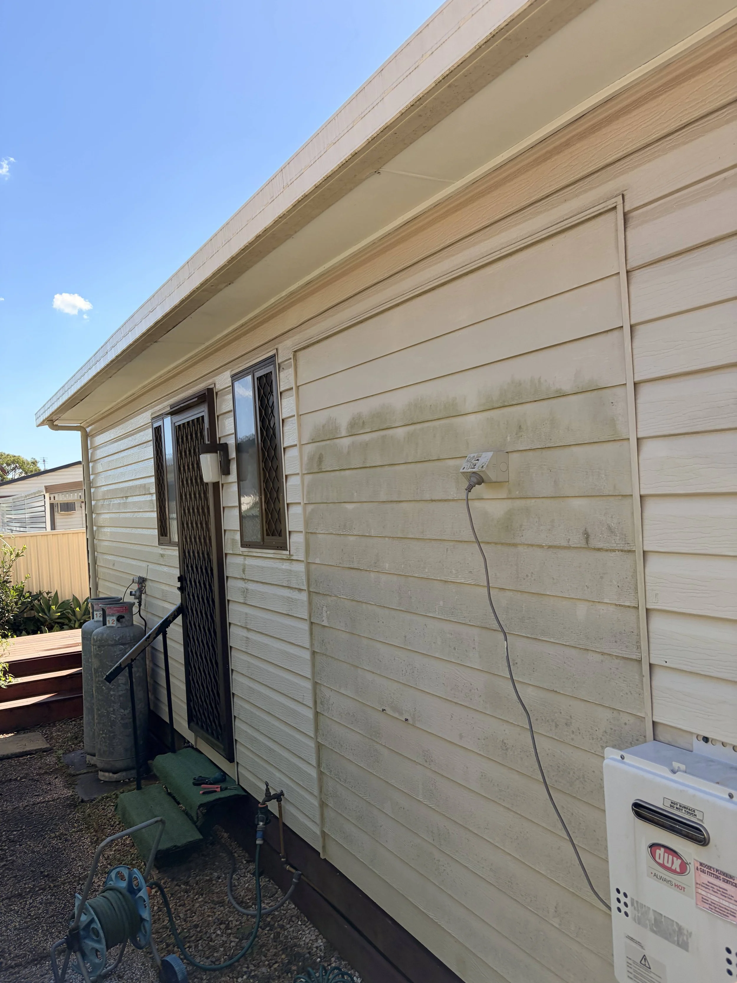 Side view of a house's exterior wall with vinyl siding, a window with security bars, a gas meter, and outdoor equipment, under a clear blue sky.