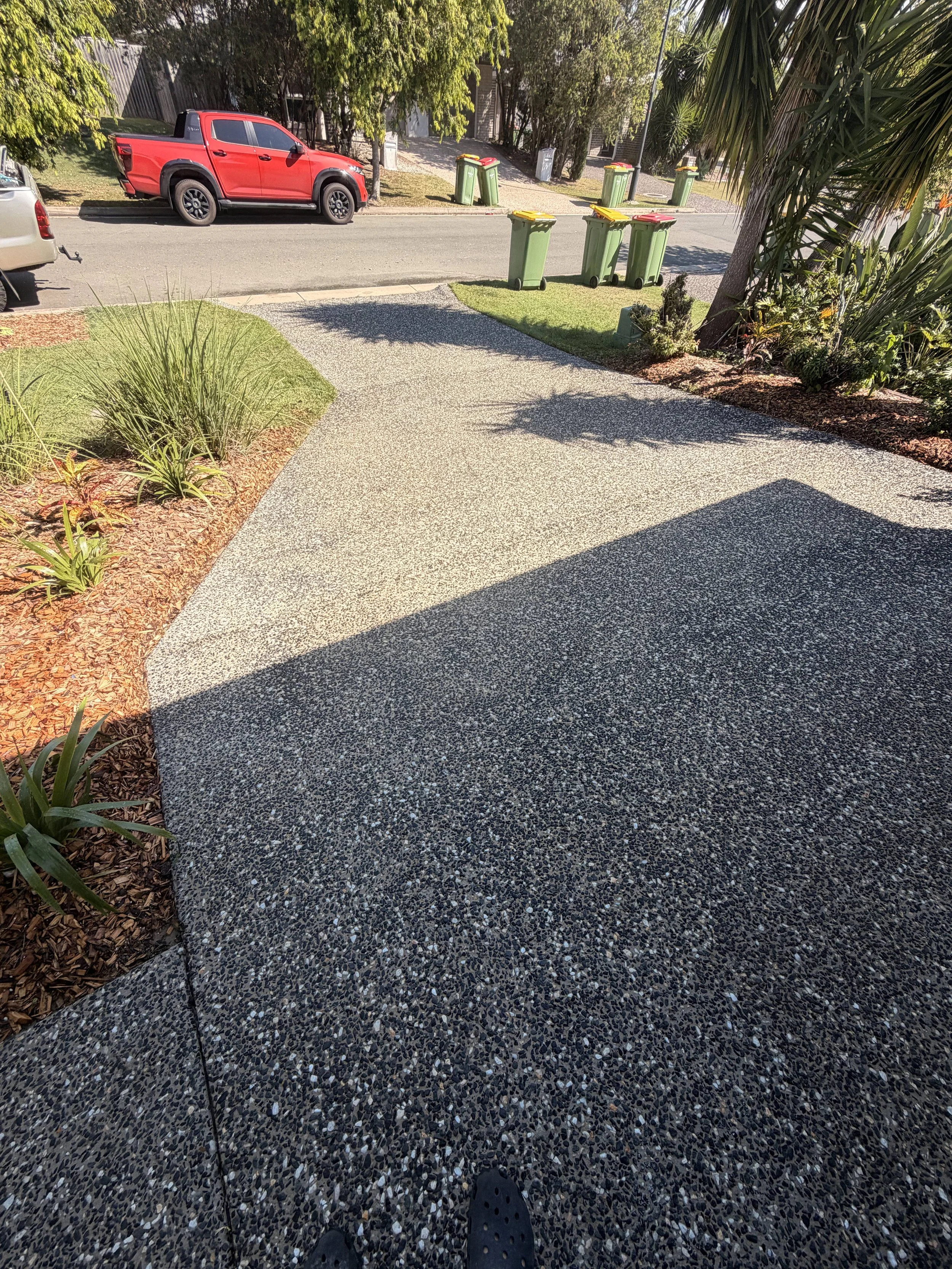 View of a concrete walkway with some plants on the left side and a garden bed on the right, with a street visible in the background. Several green and red trash bins are lined up on the sidewalk across the street. The photograph is taken from a first