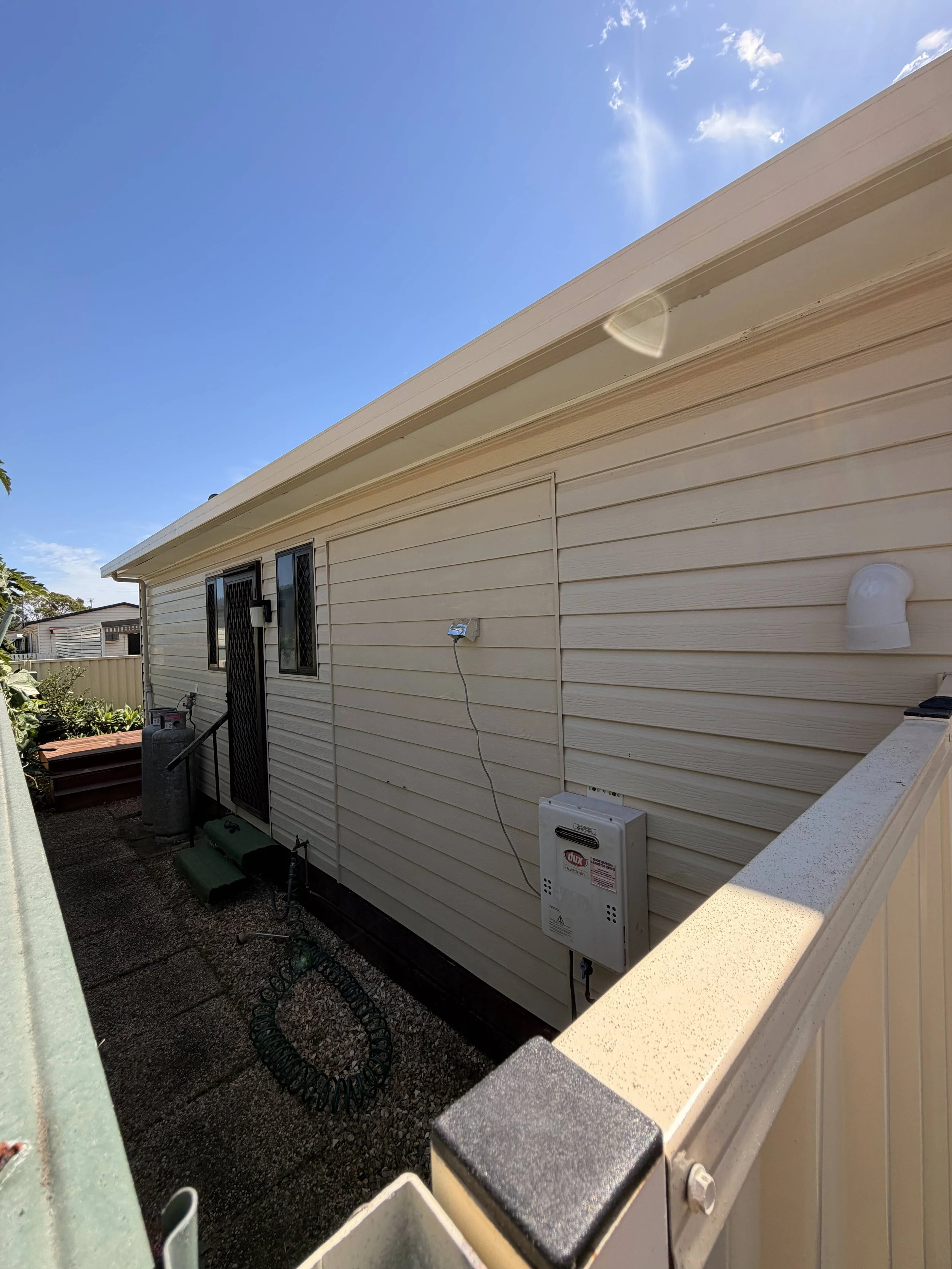Side view of a beige house exterior with black window frames, a white vent pipe, electrical box, and garden tools, under a blue sky with some clouds.