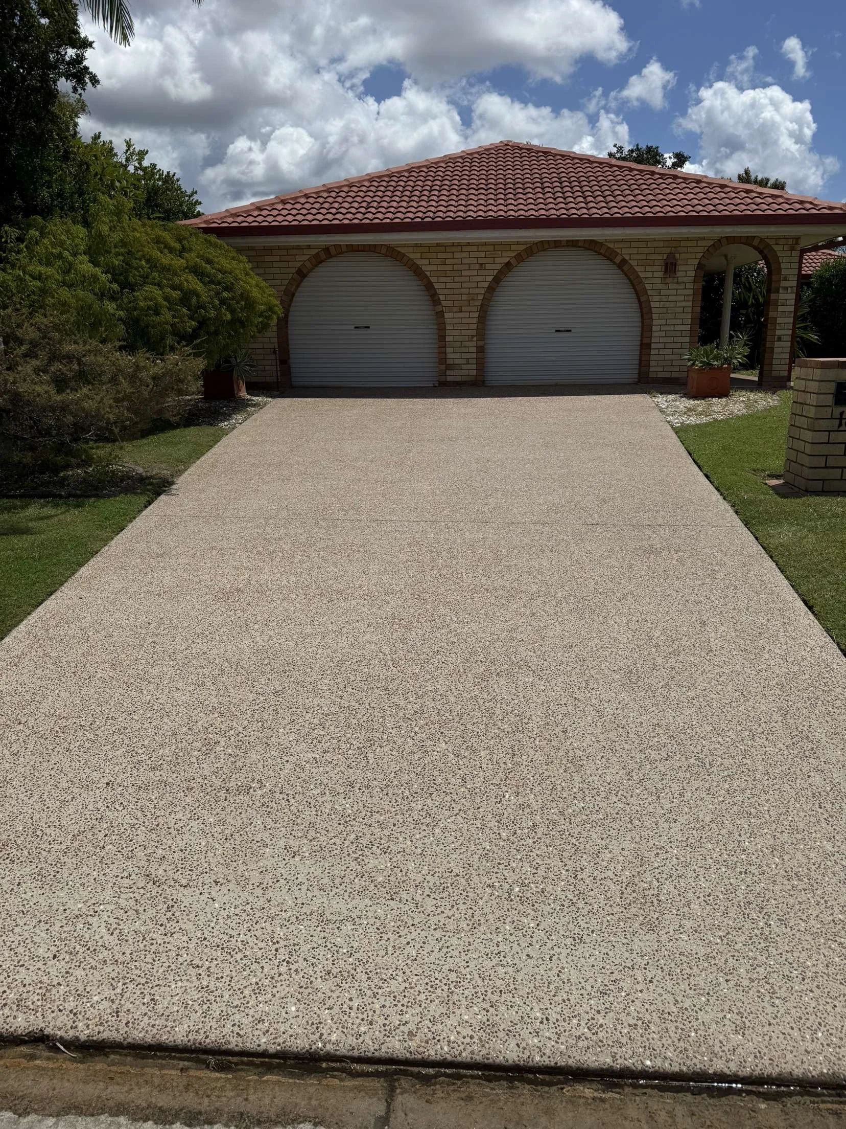 A house with a red tiled roof, brick walls, and a double garage with rolled-up white doors. There are plants in pots on either side of the garage, and a concrete driveway leading up to it. The sky is partly cloudy with a mix of clouds and blue sky.