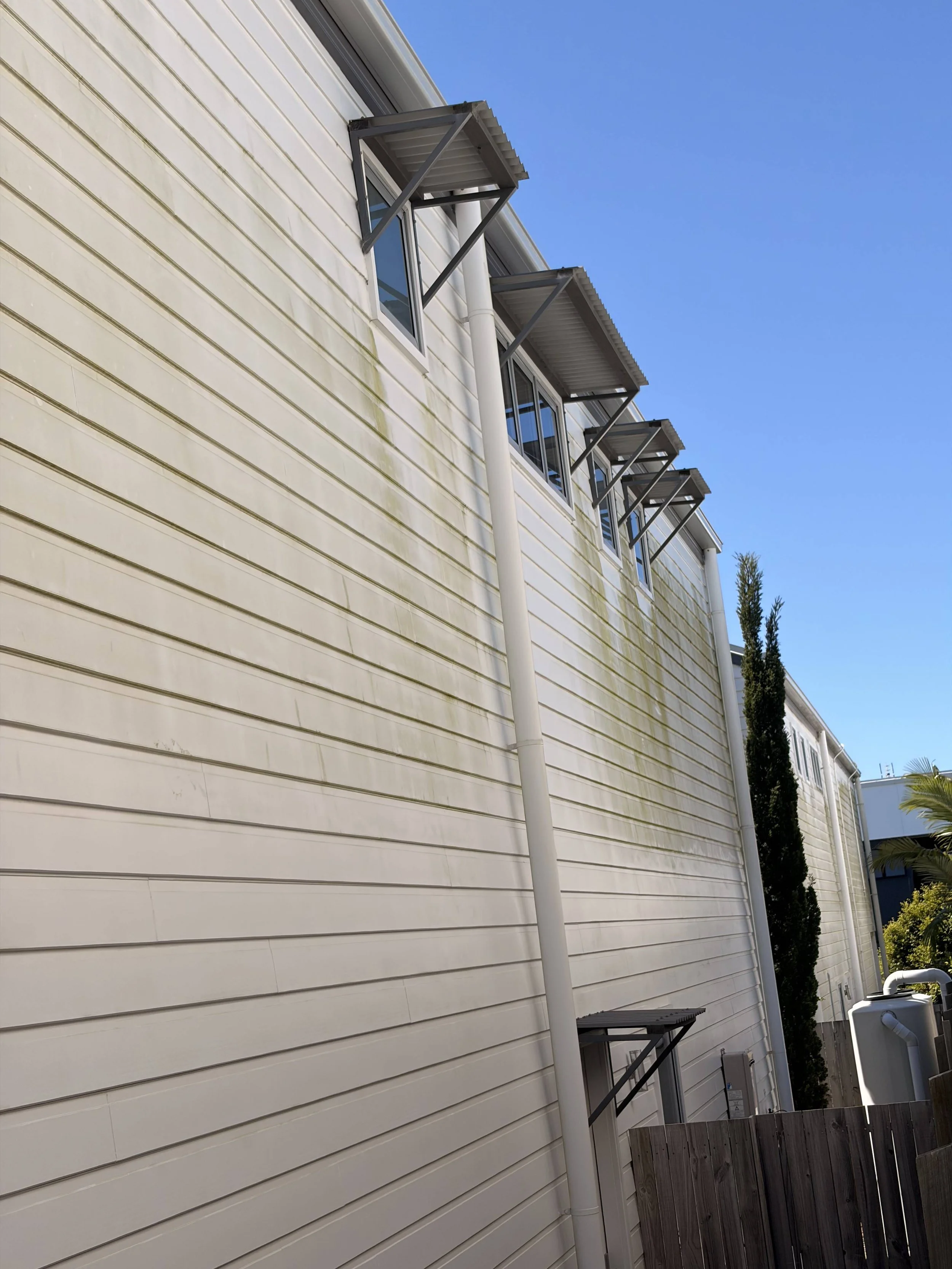 Exterior of a house with beige siding, three windows with metal awnings above them, a white drainpipe, and a wooden fence, with a blue sky and some trees in the background.