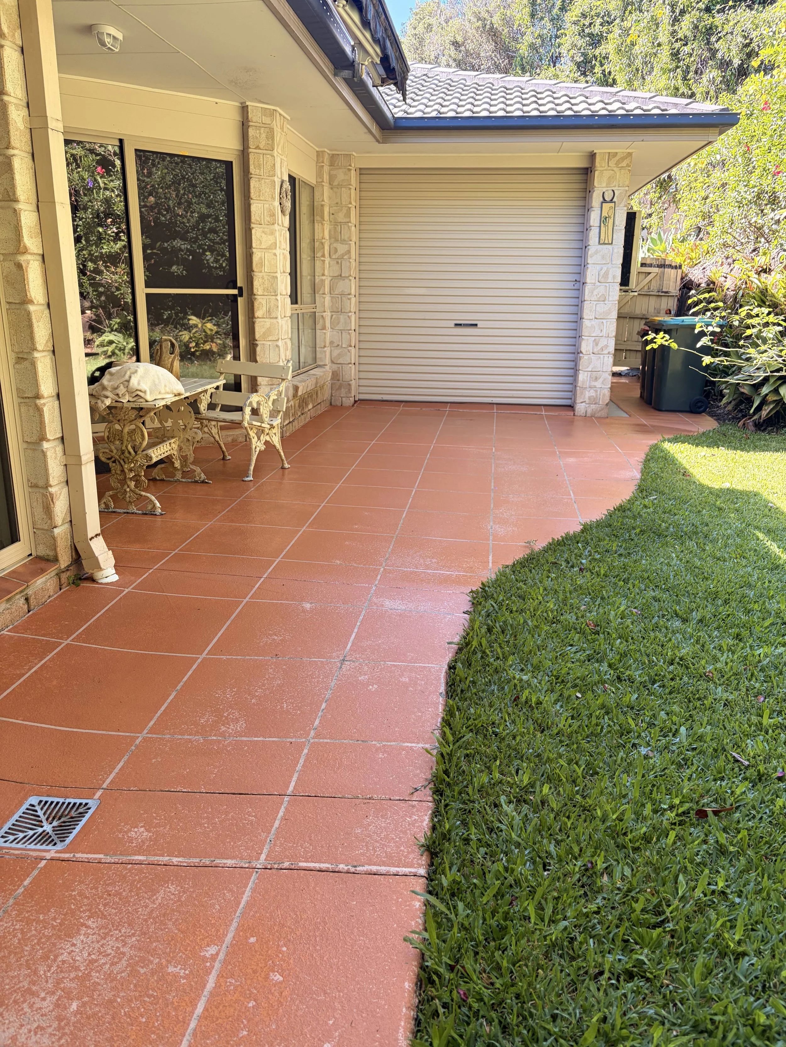 A backyard patio area with a tiled floor, a bench, a small table, a window, a garage with a shutter, and a trash bin, surrounded by green plants and trees.