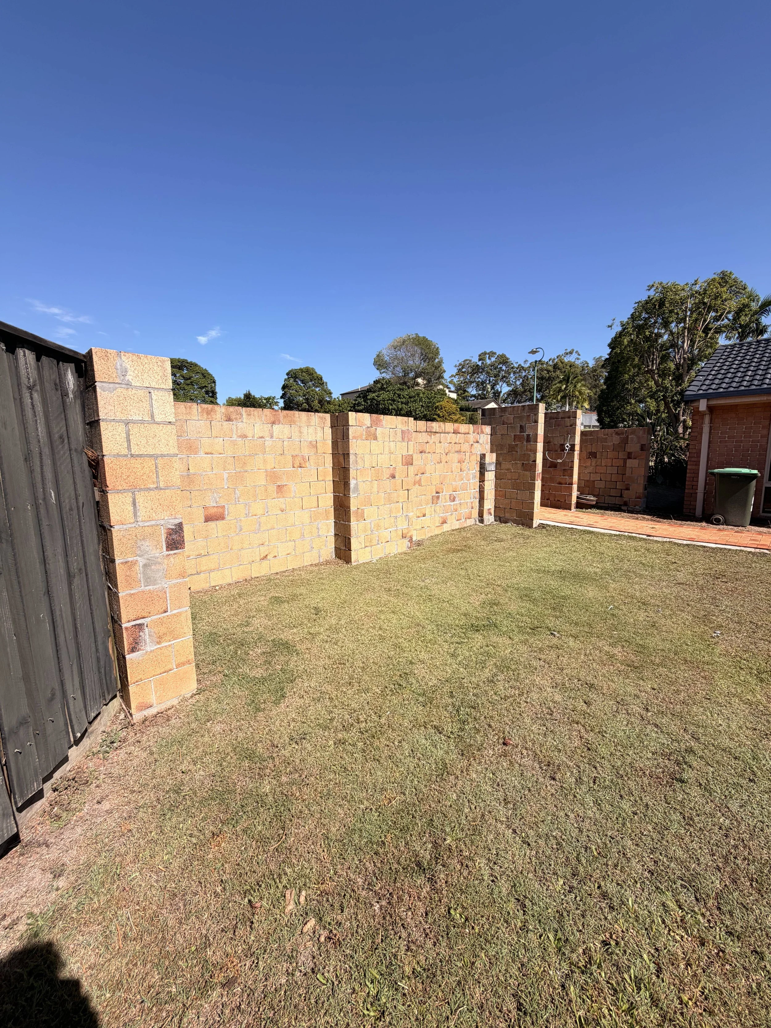 View of a backyard yard with a grass lawn, brick fence, and trees in the background under a clear blue sky.