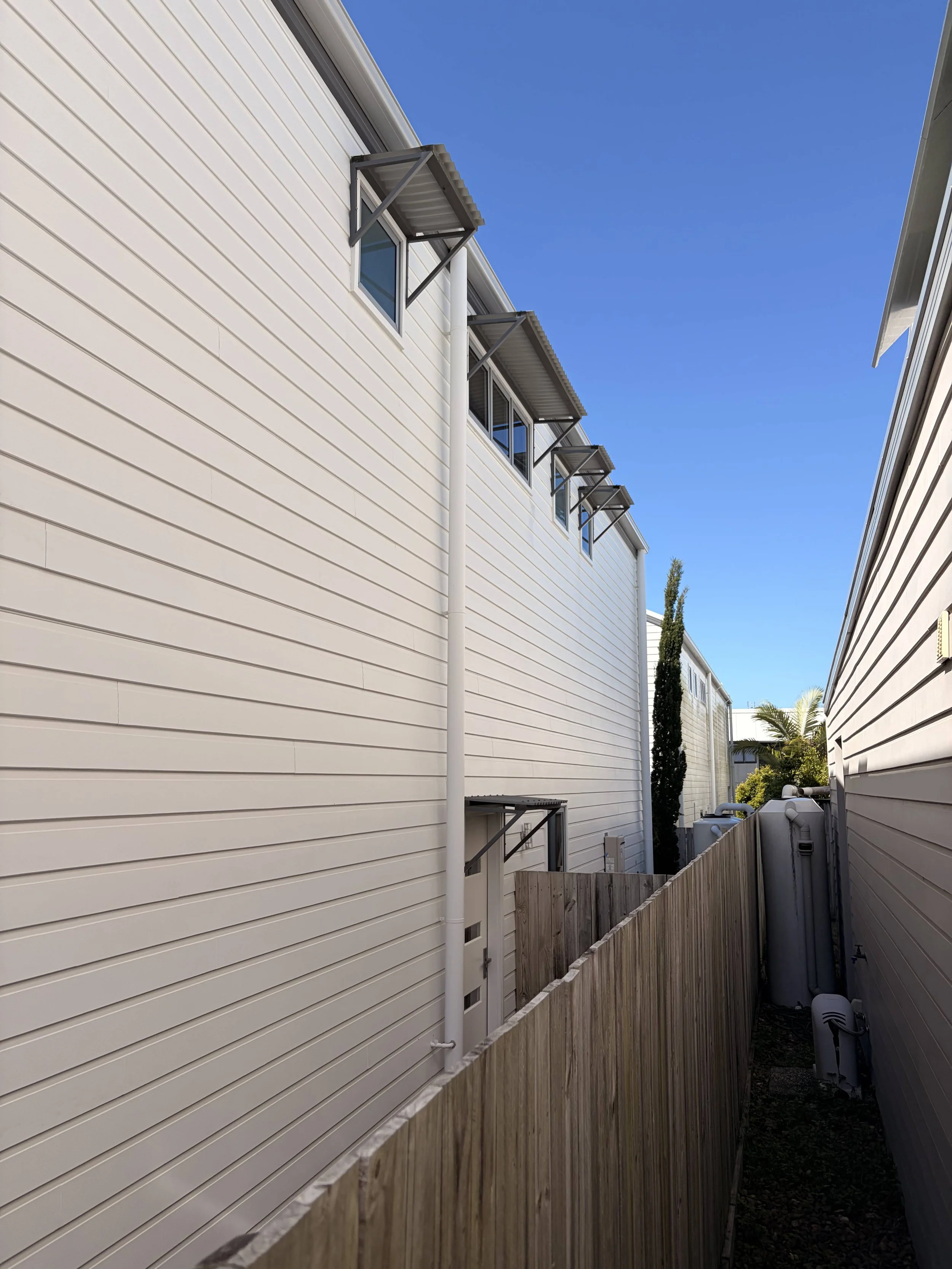 View of a narrow backyard between two white houses. The house on the left has three small awnings above windows. A tall, slim tree and some bushes are visible in the background. The sky is clear and blue.