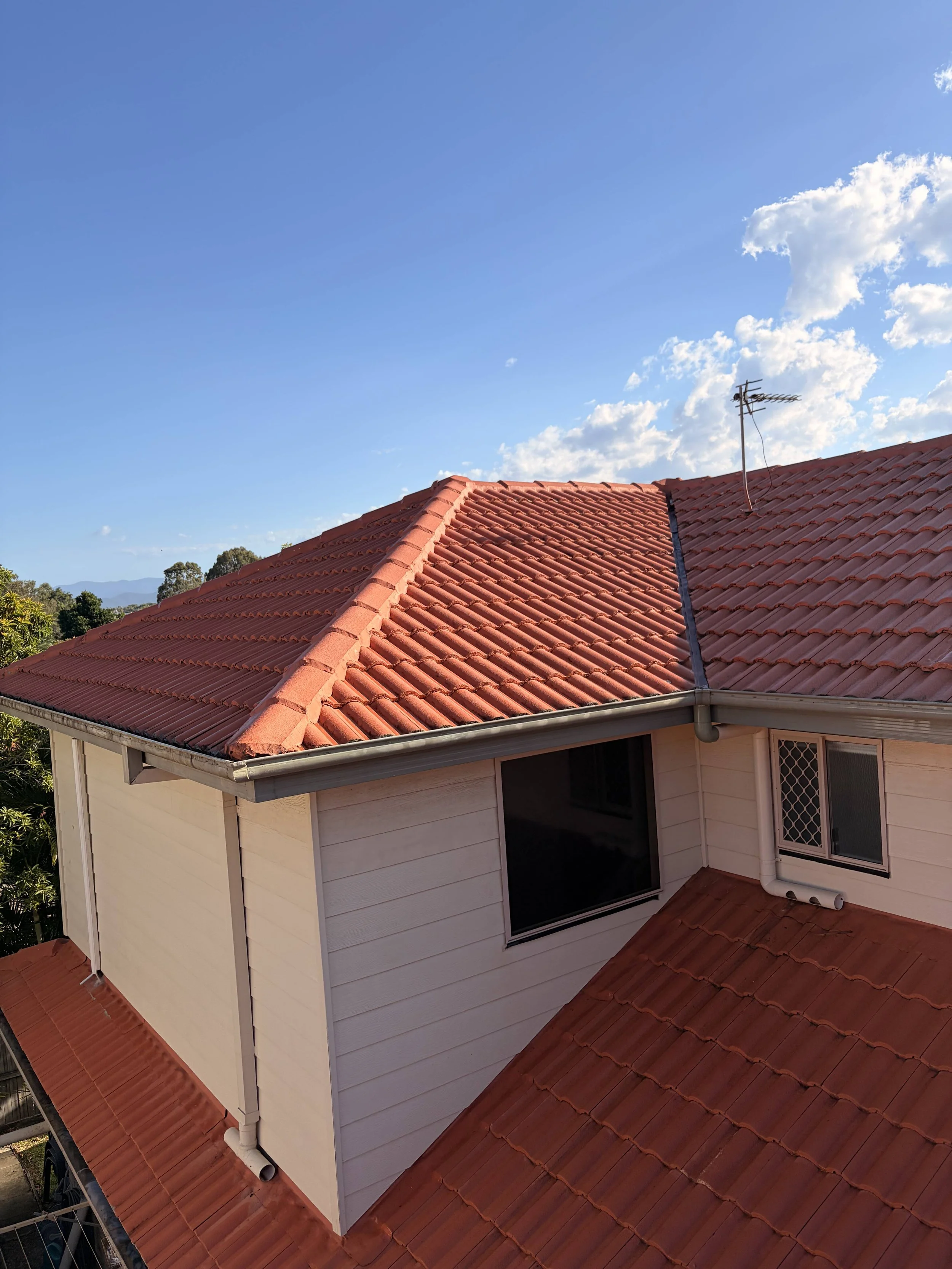 View of a house roof with red tiles, featuring a window and an antenna, under a blue sky with scattered clouds. Trees are visible in the background.