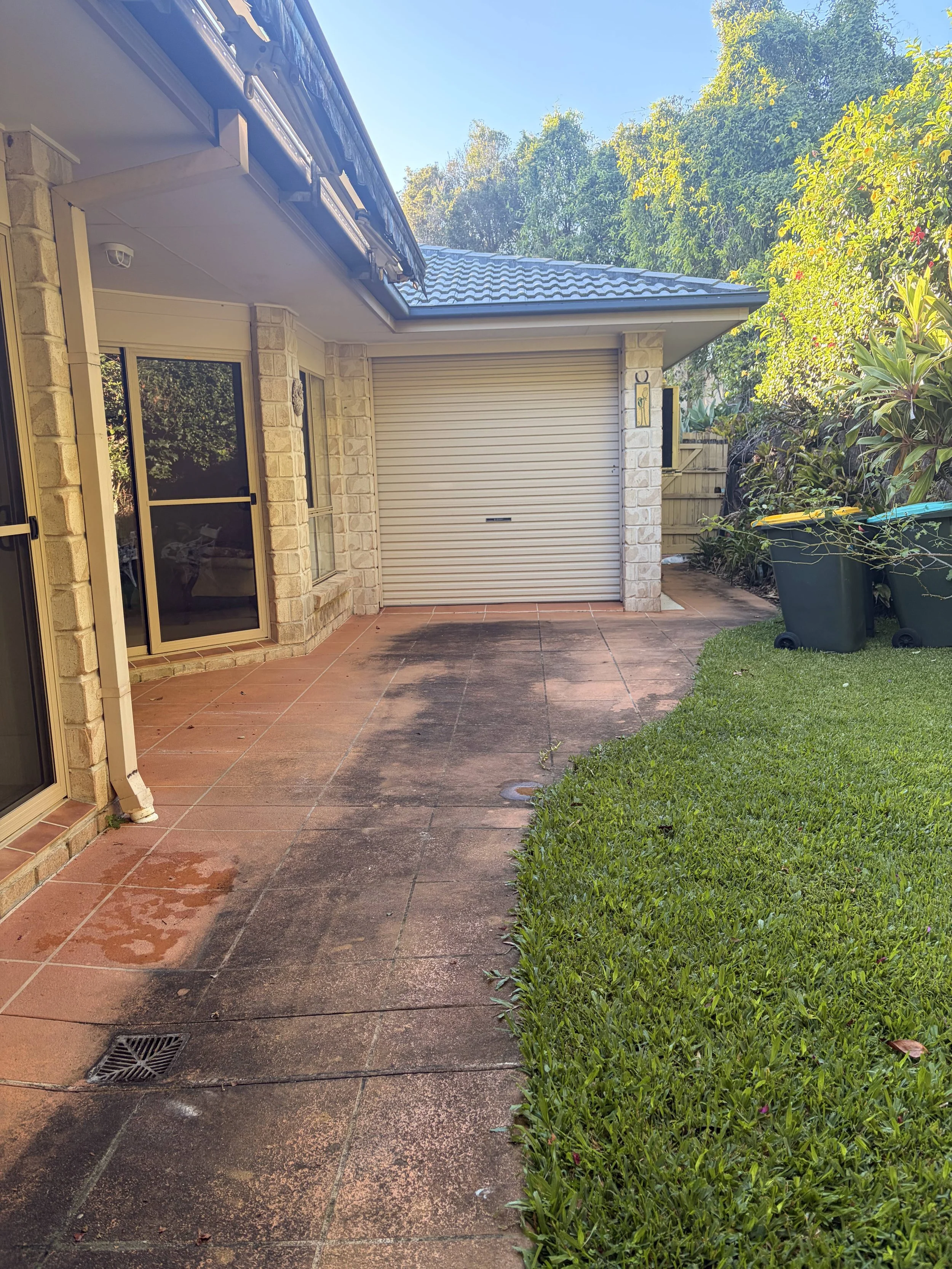 Back patio area of a house with a tiled floor, brick walls, a roller door, sliding glass door, sidewalk, green grass, and plants with trees in the background.