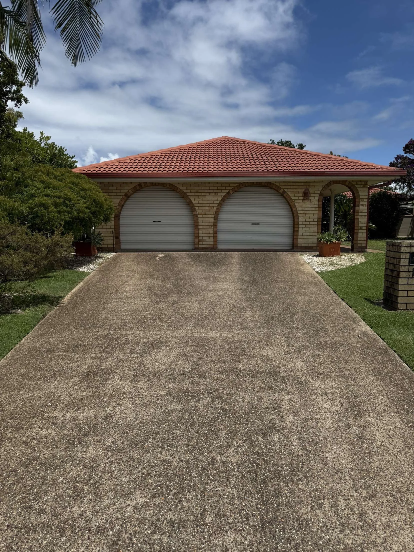 View of a concrete driveway leading to a garage with two white rolling doors under a red-tiled roof. The garage is surrounded by green bushes and trees, with a partly cloudy blue sky overhead.