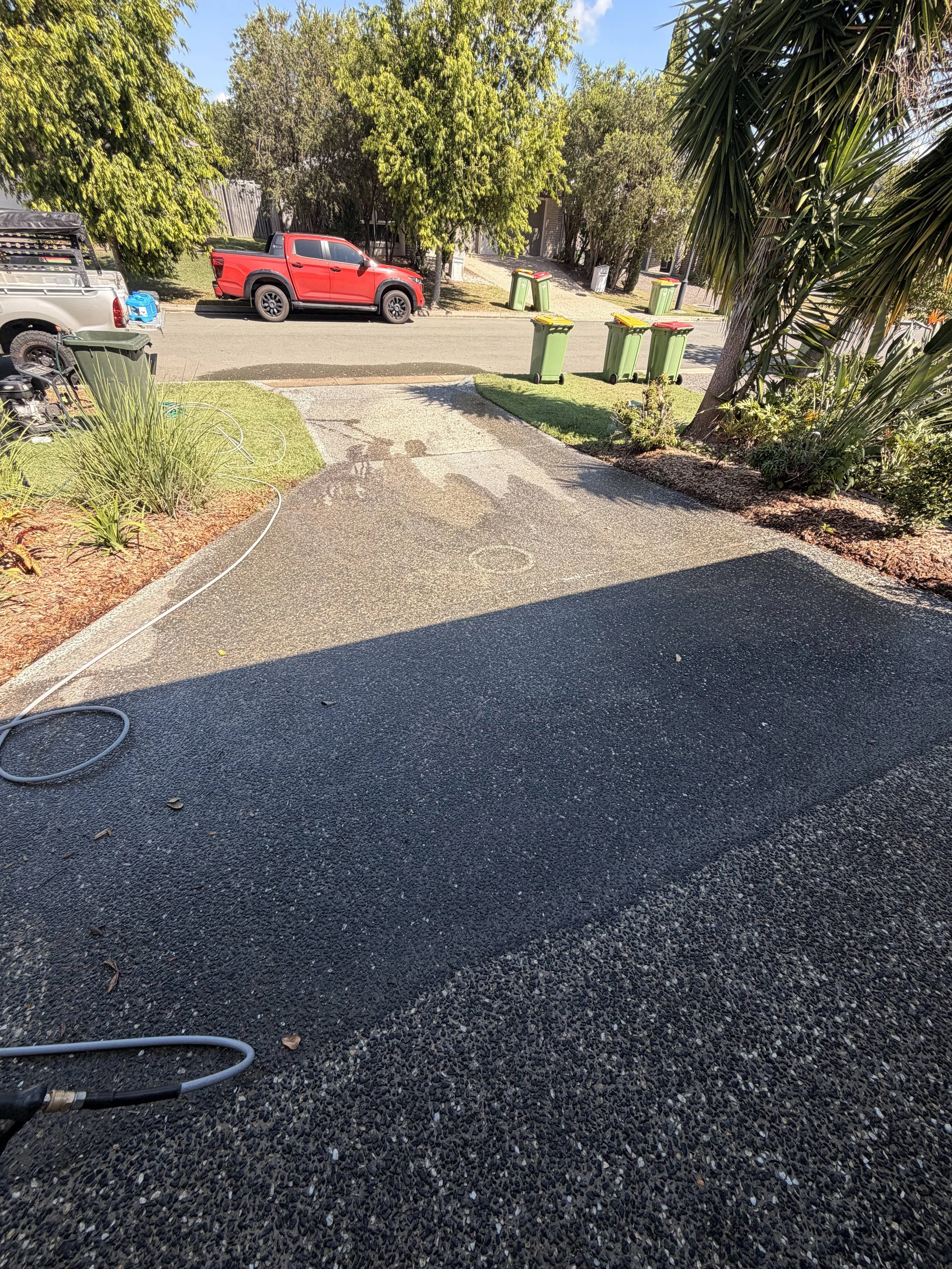 A driveway with a garden hose on it, a concrete planter, and bushes on both sides, leading to a street with a red pickup truck parked on the side and five green garbage bins with yellow and red lids in front of the street. There are trees and a sidew