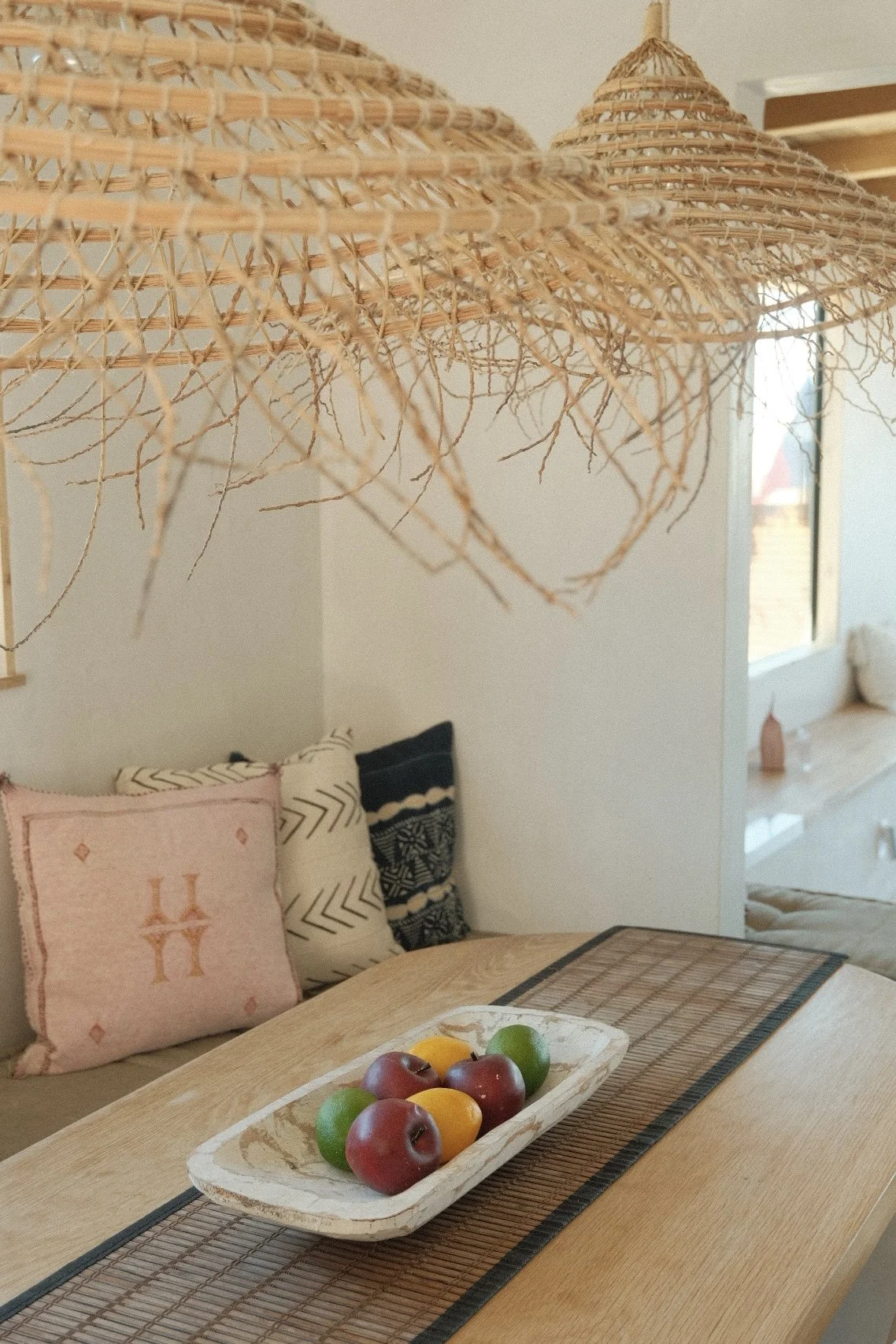 A wooden dining table with a rectangular woven placemat and a stone bowl filled with red and yellow apples and green limes. In the background, embroidered cushions are on a bench against the wall, with a large wicker light fixture hanging above.