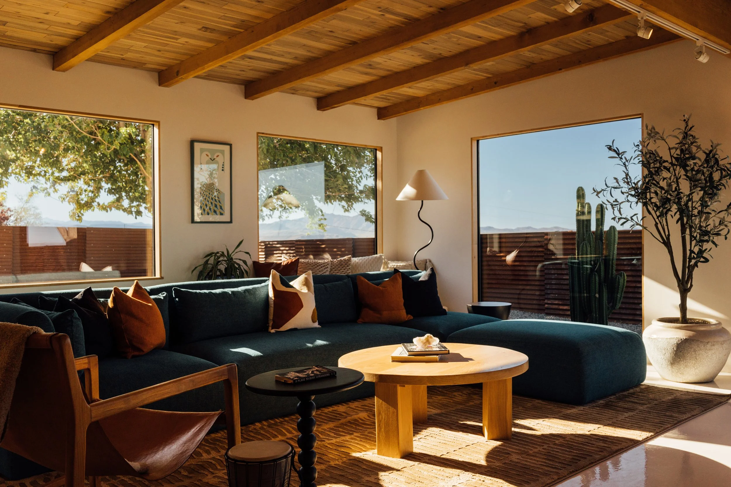 Living room with wooden vaulted ceiling, large windows showing outdoor trees and mountains, navy sofa with colorful pillows, wooden coffee table, artwork on wall, white floor lamp, potted plant, cactus, and a patterned rug.