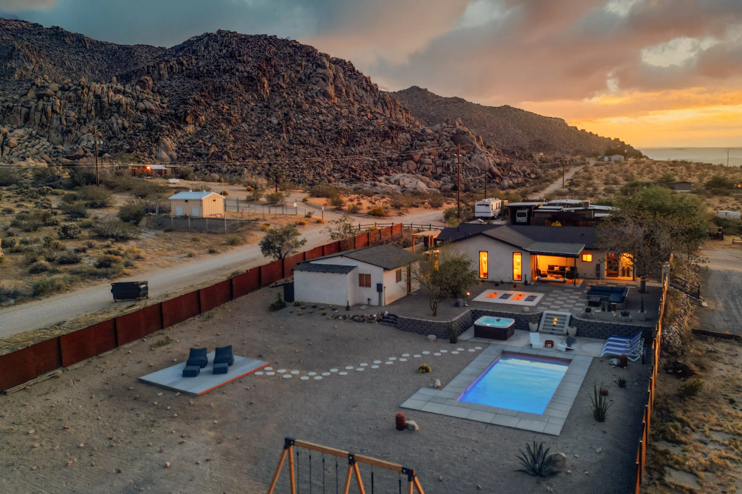 A residential backyard with a swimming pool, outdoor seating area, surrounded by desert landscape with boulders in the background during sunset.