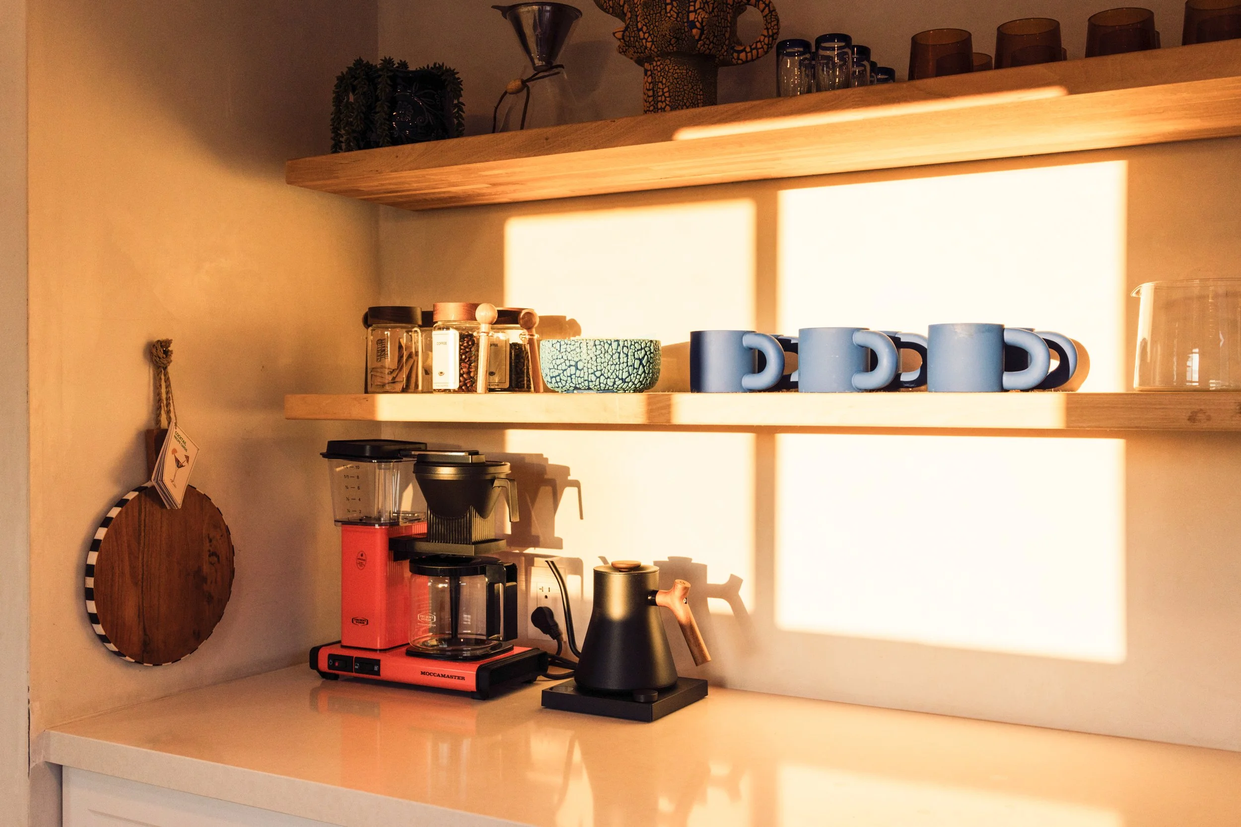 Kitchen shelf with coffee cups, jars, and decorative items illuminated by warm sunlight.