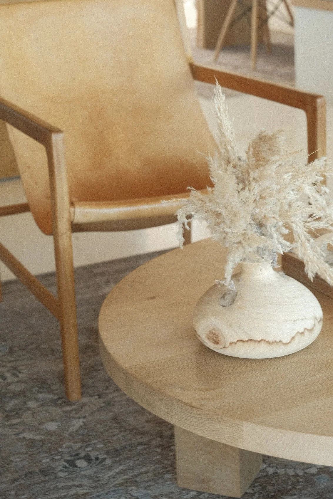 A beige armchair with wooden armrests next to a round wooden table with a beige vase holding dried pampas grass.