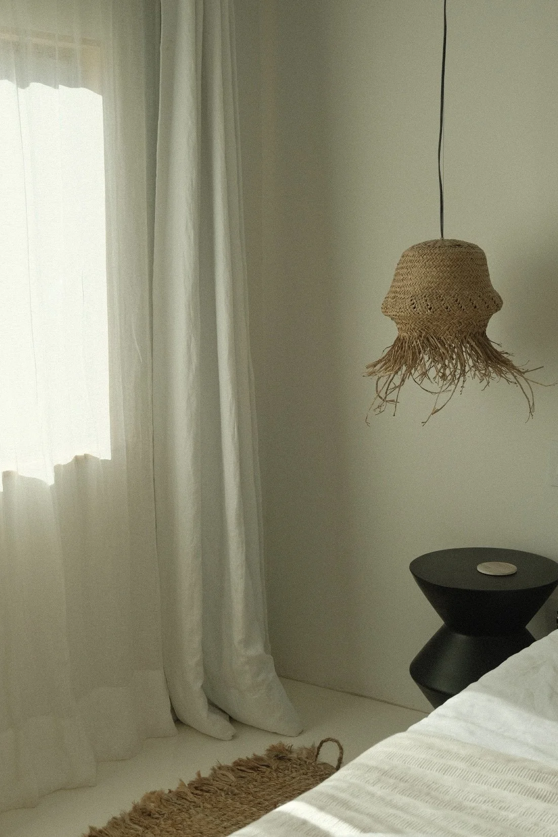 A bedroom corner with a window covered by white curtains, a hanging woven lampshade, a black bedside table with a small round object, part of a bed with white bedding, and a brown rug.