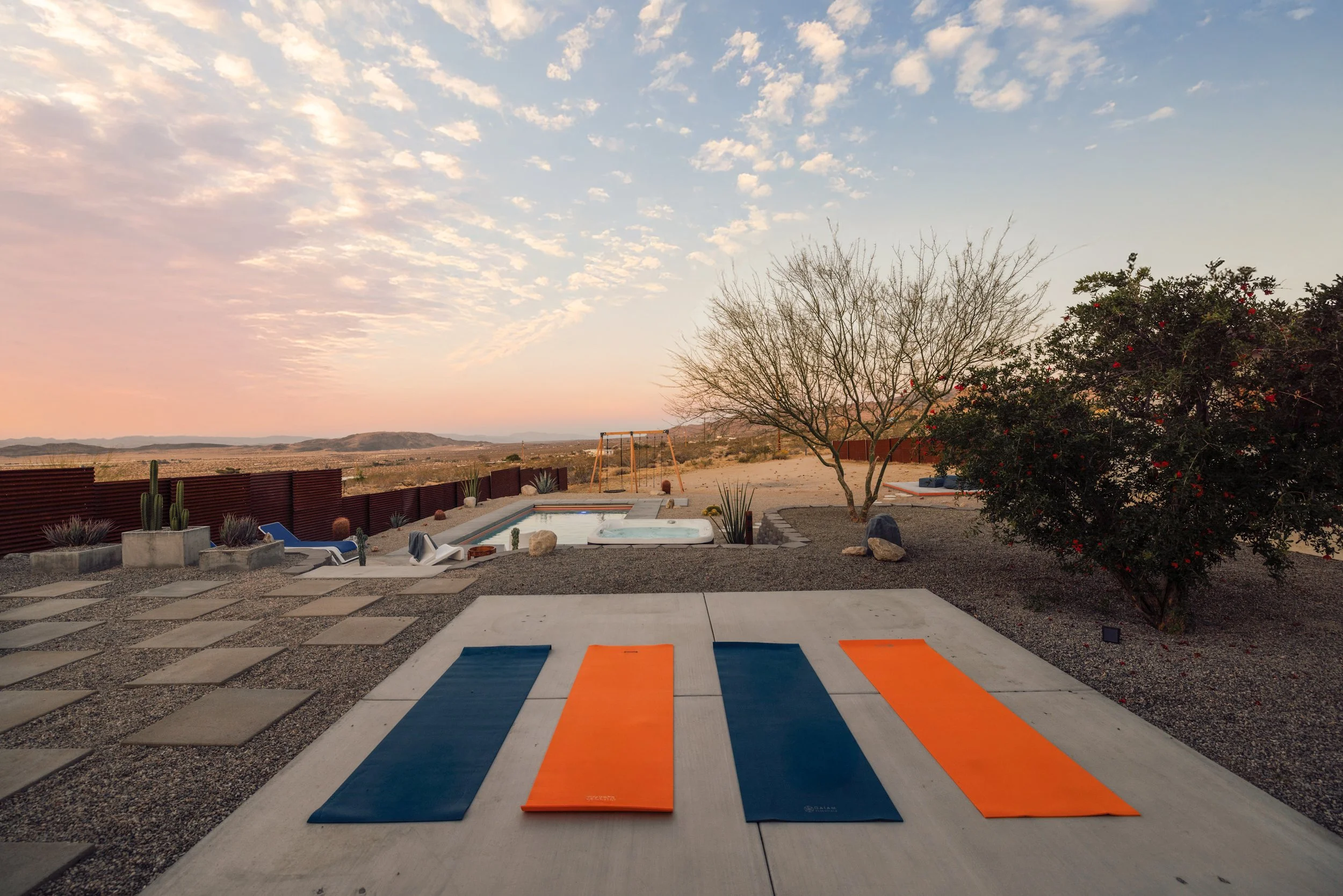 A backyard with a pool, desert landscape, and mountains in the distance. The sky is partly cloudy during sunset. There are yoga mats laid out on concrete and pebble ground, and a tree with sparse branches and another bush with red flowers.