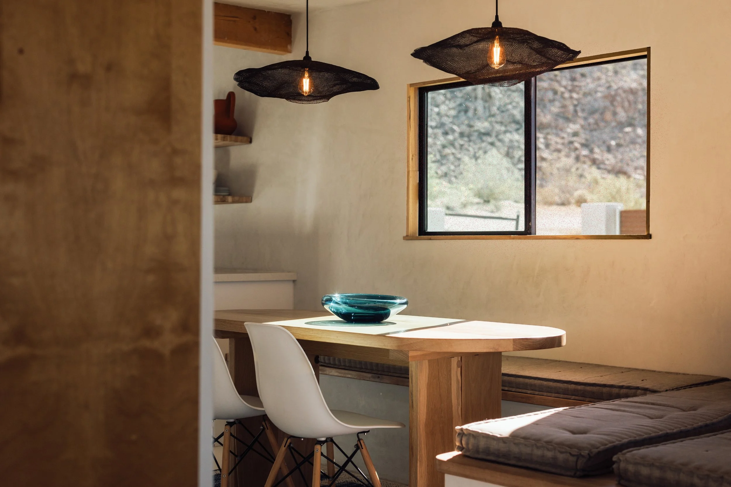A wooden dining table with a decorative glass bowl on top, surrounded by white chairs, in a room with a window and two black pendant lights hanging from the ceiling.