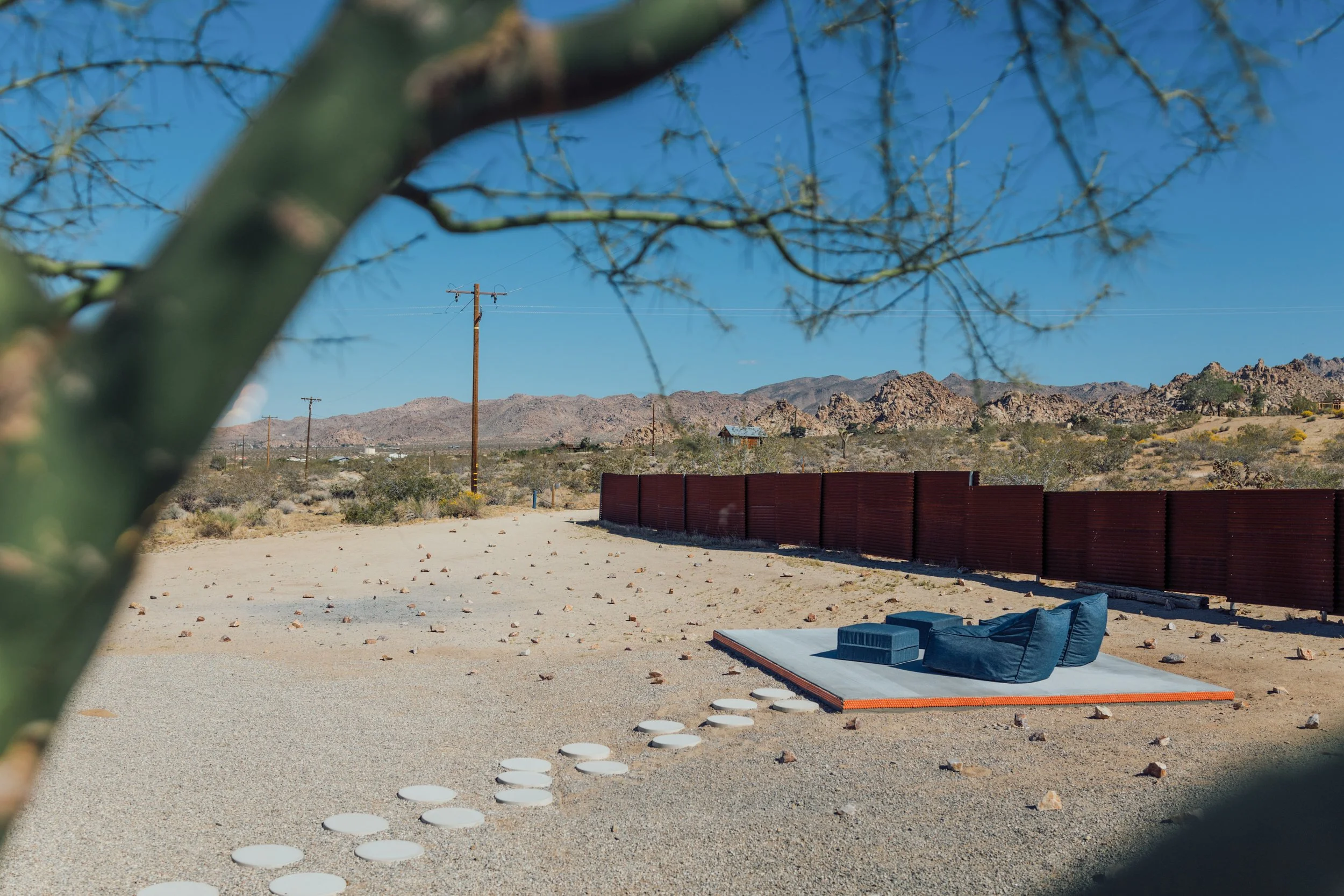 View of a desert landscape with mountains in the background, utility poles, and a red fence. In the foreground, there is a gravelly surface with outdoor furniture and circular stepping stones.