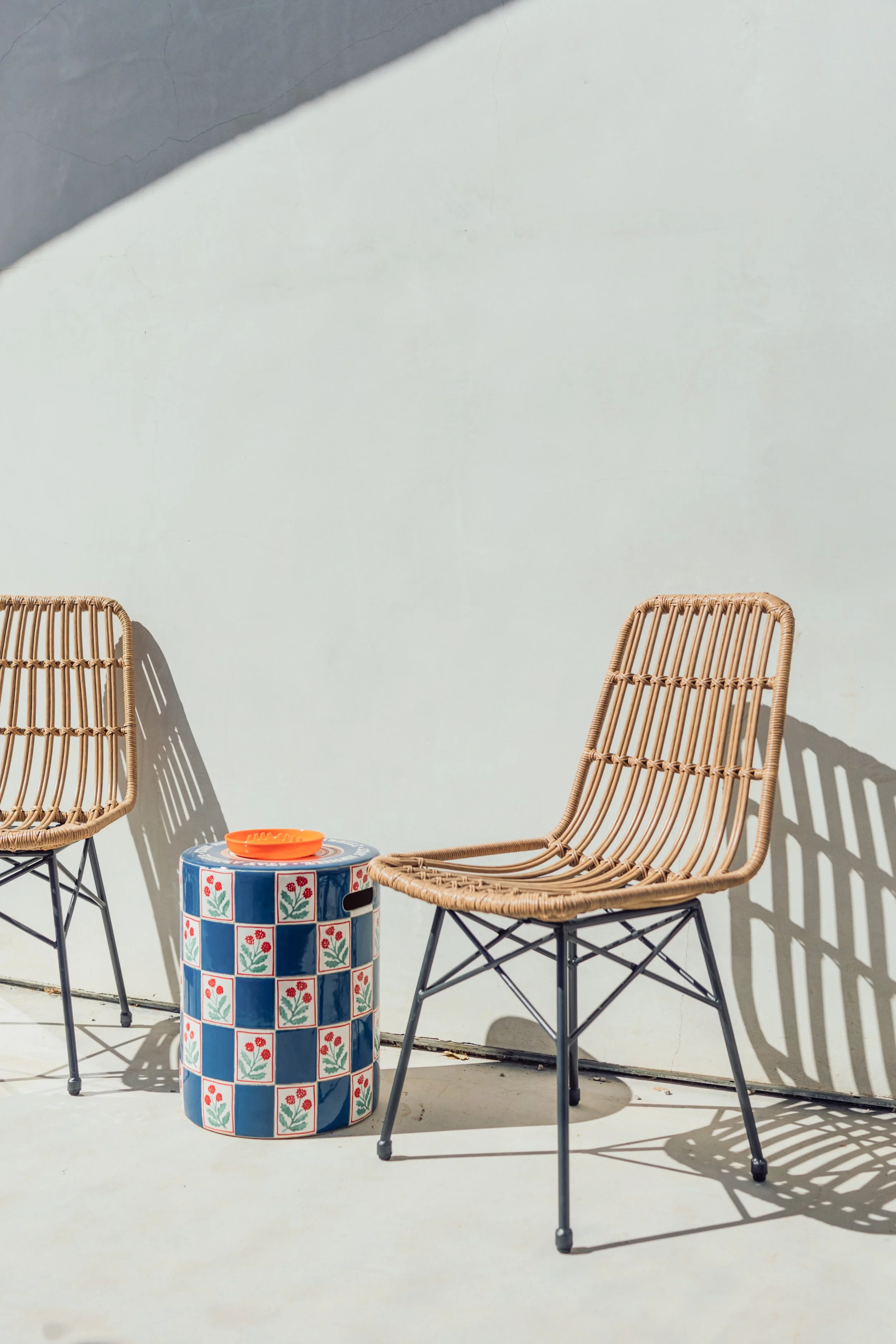 Two rattan chairs with black metal legs are positioned against a light-colored wall. Between them is a round ceramic table with a floral pattern, topped with an orange bowl. Shadows of the chairs are cast on the ground.