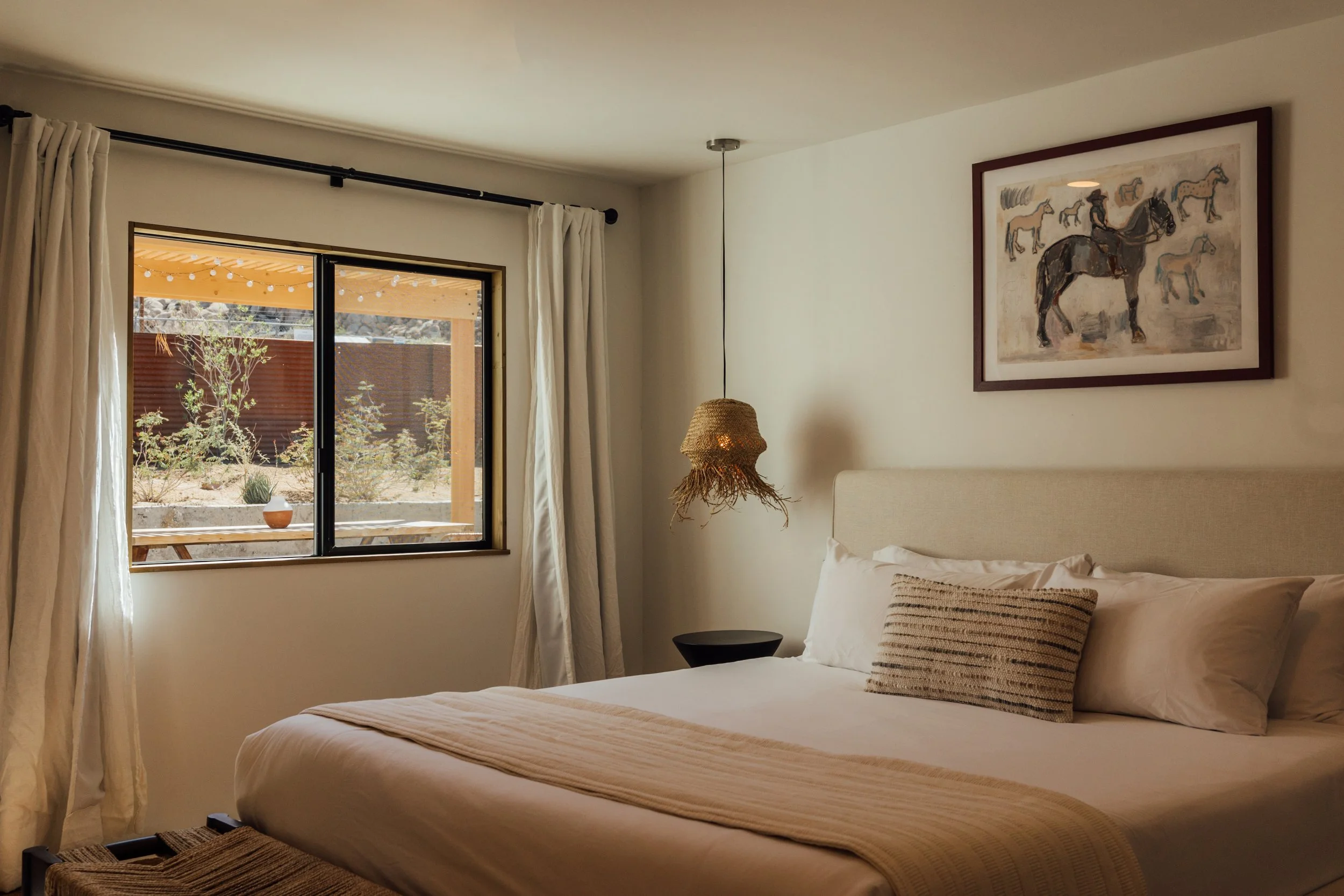A bedroom with a large bed, white bedding, and a textured throw pillow. There is a window with white curtains, overlooking a patio with string lights and desert plants. A framed painting of horses hangs on the wall, and a woven hanging light fixture is beside the bed.