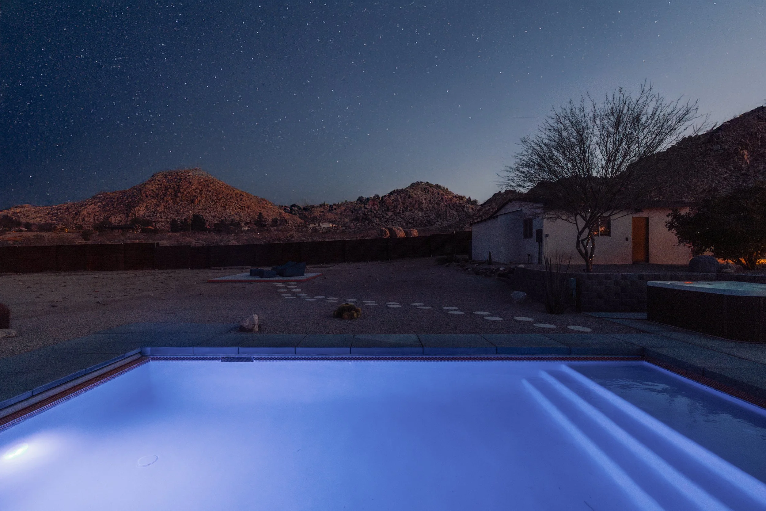 Nighttime scene of an outdoor swimming pool illuminated with blue lights, surrounded by a desert landscape with mountains and a house, under a starry sky with few clouds.