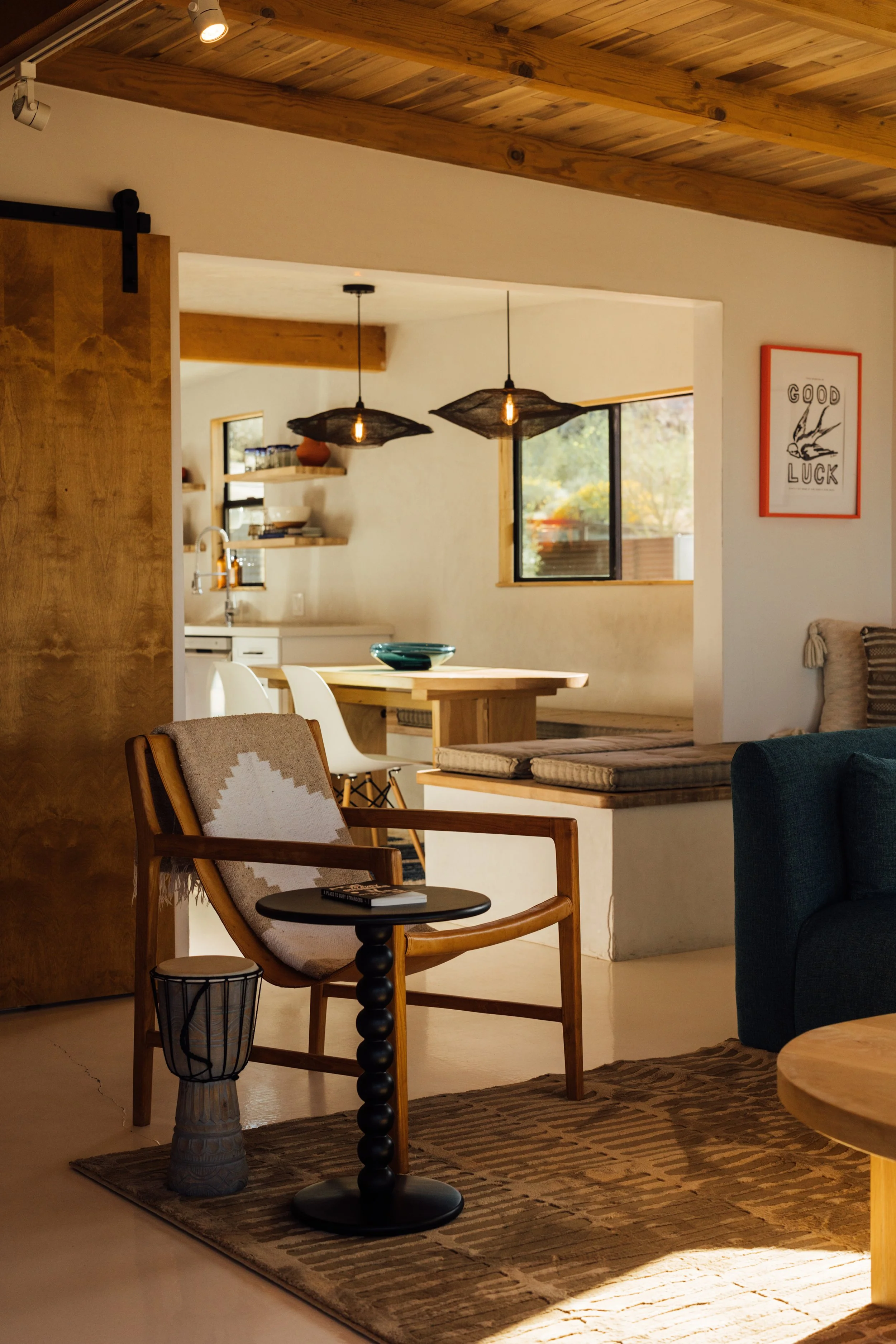 A cozy interior of a living room with a mix of modern and rustic decor. Features include a wooden armchair with a beige and white patterned pillow, a small black side table with a book, a tall woven basket, a dark green sofa, a wooden dining table with white chairs, and decorative pendant lights hanging from the ceiling. The room has natural light coming through a window with a view of outdoor trees and a framed print on the wall that reads 'Good Luck' with a bird illustration.