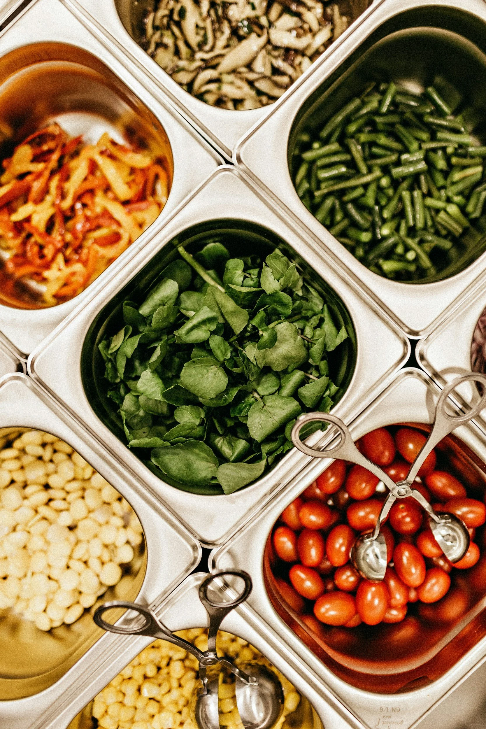 A self-serve salad bar with containers holding cherry tomatoes, chopped green beans, chopped mushrooms, shredded carrots, mixed vegetables, and leafy greens.