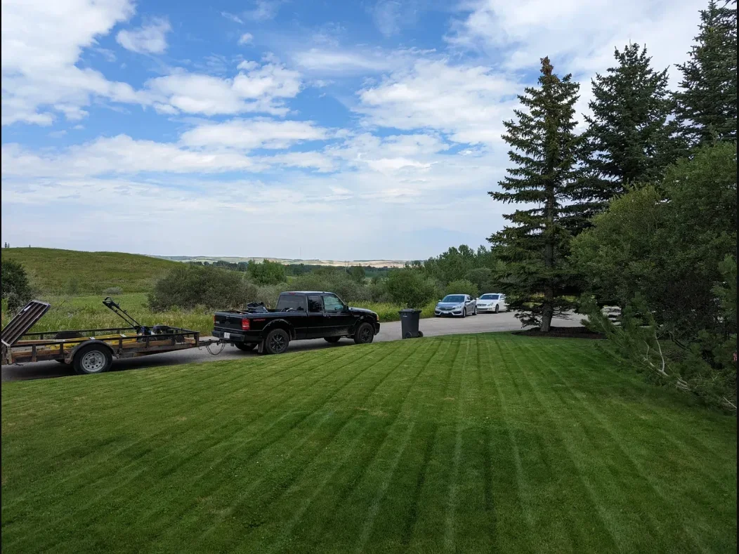 A black pickup truck parked on a roadside with a trailer attached, backed by a neatly mowed lawn and several trees, with a few cars visible on the road and a partly cloudy sky overhead.