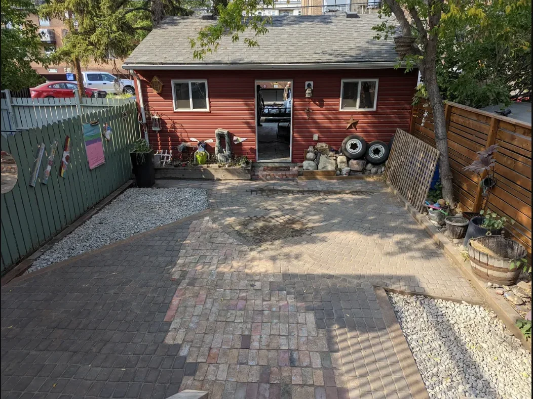A backyard with a paved patio, a red shed, a wooden fence, and decorative elements such as tires, rocks, a star, and potted plants. There are trees providing shade and a gravel area beside the patio.