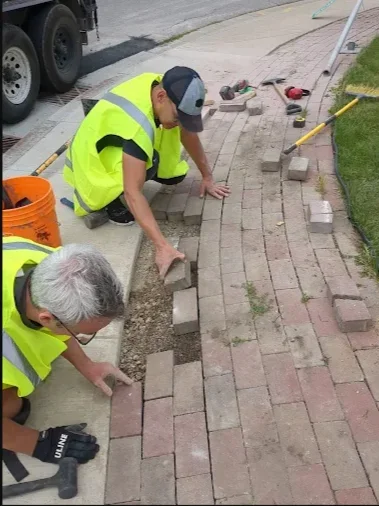 Two workers in yellow safety vests installing bricks beside a sidewalk and lawn