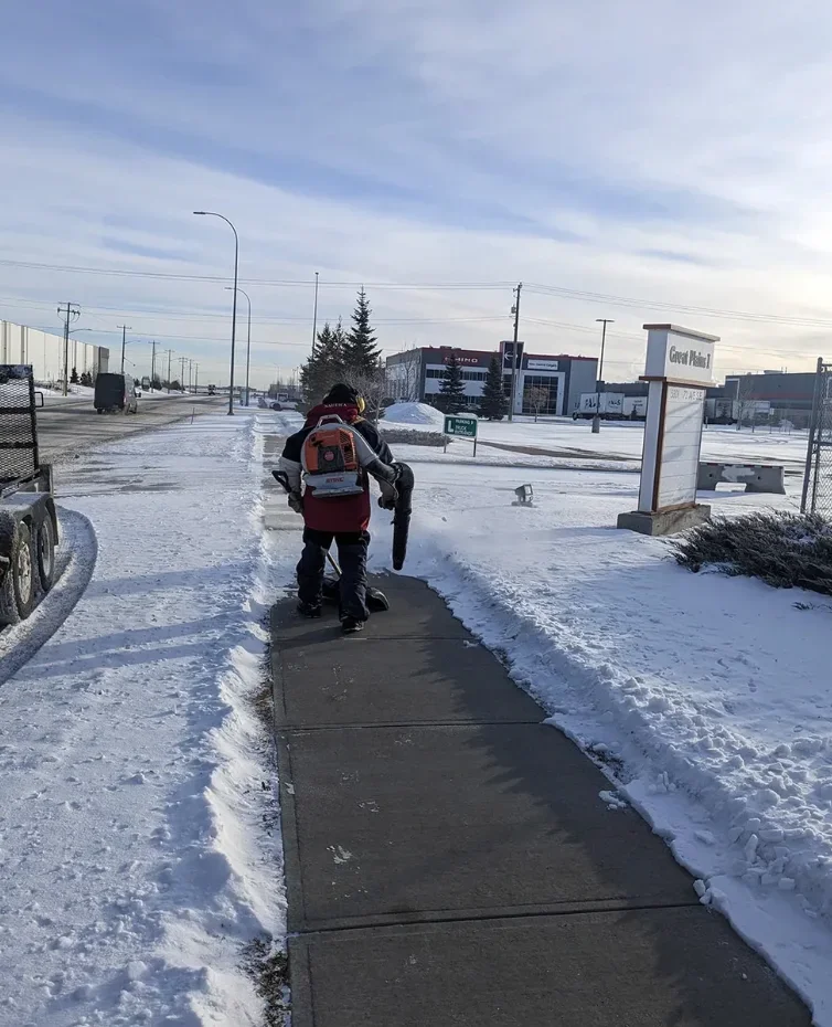 A person walking on a snowy sidewalk and removing snow with a blower and shovel.