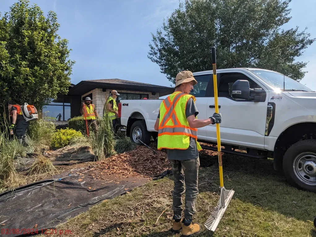 A man in a reflective safety vest and camouflage pants is holding a rake and standing on the grass, while other workers in similar vests and helmets are near a white pickup truck on a lawn with trees and a small building in the background.