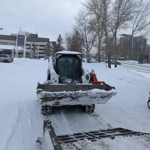 A skid steer clearing snow from a sidewalk beside a plaza in winter.