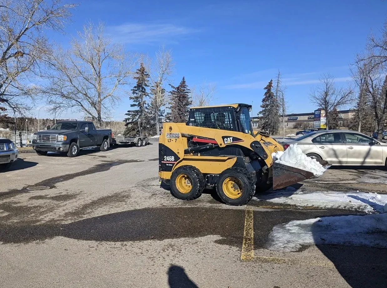 A yellow skid-steer loader removing snow from a parking lot, with parked cars and leafless trees in the background on a clear, sunny winter day.