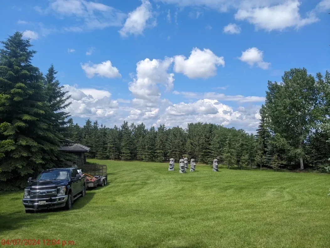 A spacious grassy lawn with tall trees on the sides and a line of five moai statues in the background. A black pickup truck with a trailer is parked on the left side near a small building. The sky is partly cloudy with white fluffy clouds.